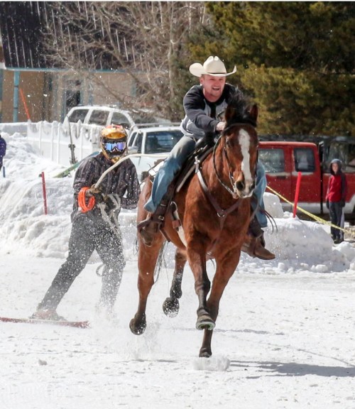 A cowboy on a horse races through snow while being pulled by a person on skis, creating a dynamic winter scene.