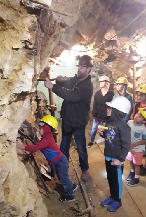 A group of people, including children, explore a mine, wearing hard hats and safety gear while observing a demonstration.