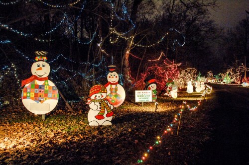 Colorful holiday decorations line a path, featuring snowmen and twinkling lights in a nighttime setting.