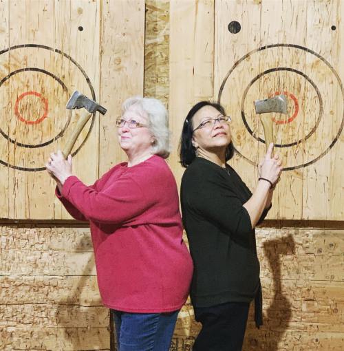 Two women stand back-to-back, each holding an axe, in front of wooden targets with red circles.
