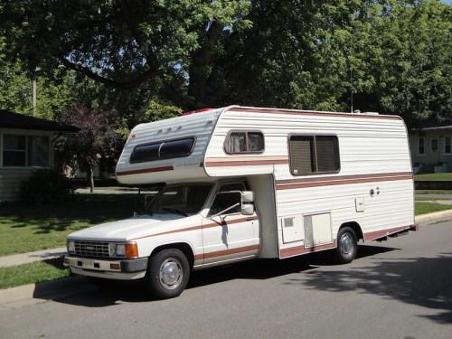 A white and brown camper van parked on a residential street, surrounded by green trees and houses.