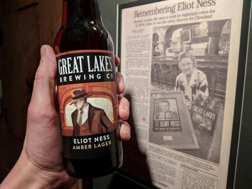 A hand holds a bottle of Great Lakes Eliot Ness Amber Lager next to a framed newspaper article about Eliot Ness.