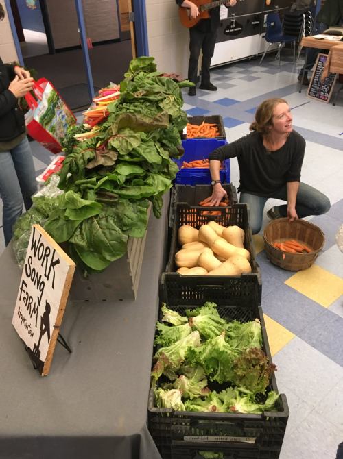 A display of fresh vegetables, including greens and carrots, with a woman kneeling beside baskets in a community setting.