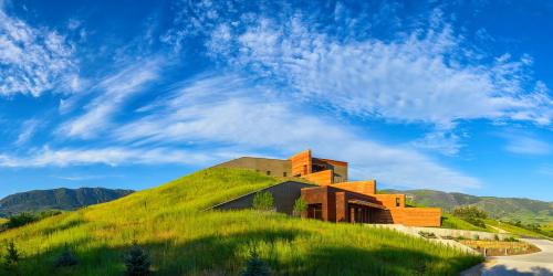 A modern building on a grassy hill, surrounded by mountains and a blue sky with wispy clouds.