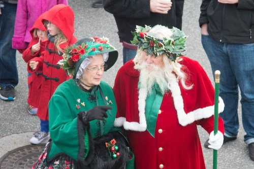 A woman in a green dress and a man in a red Santa suit chat, both adorned with festive decorations.