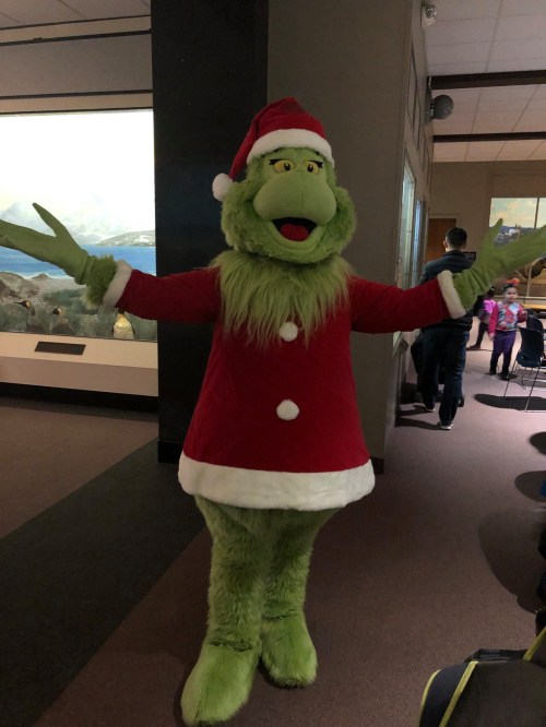 A person in a green Grinch costume wearing a red Santa outfit, standing with arms outstretched in a museum setting.