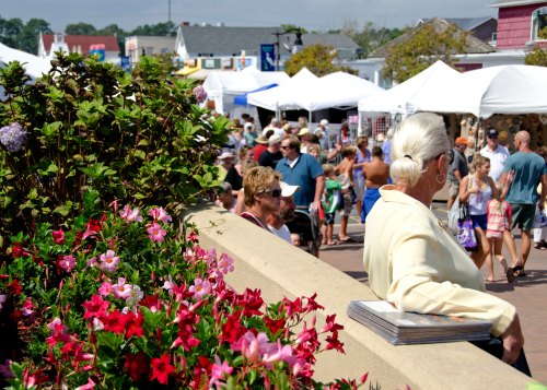 A bustling outdoor market scene with colorful flowers in the foreground and crowds browsing tents in the background.