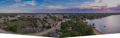 Aerial view of a lakeside town with greenery, a crowd gathering, and a serene lake under a colorful sky.