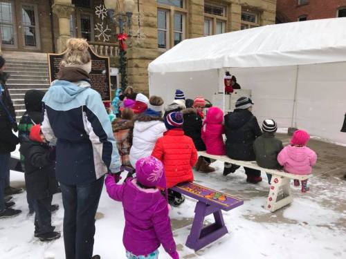 A group of children in colorful winter coats watch a performance under a tent in a snowy outdoor setting.