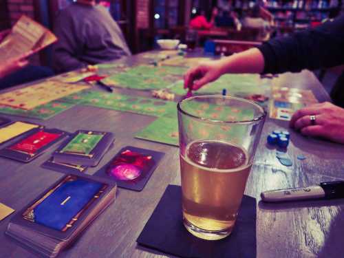 A tabletop game scene with cards, game pieces, and a glass of beer on a wooden table in a cozy setting.