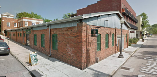 A brick building with green windows on a street corner, featuring a sign and nearby parked cars.