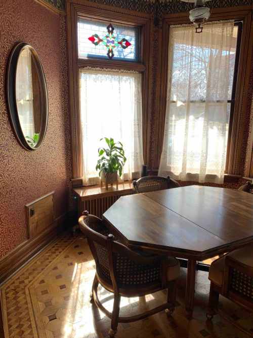 A cozy dining area with a wooden table, chairs, and large windows adorned with lace curtains and a decorative mirror.