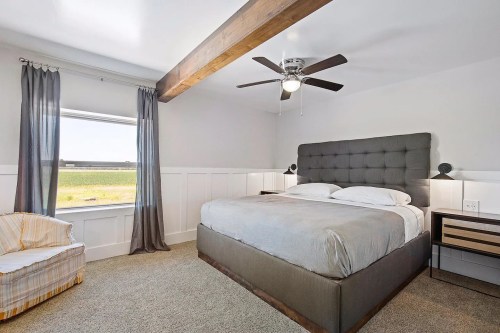 A modern bedroom featuring a large bed, ceiling fan, and window with a view, complemented by gray curtains and a cozy chair.