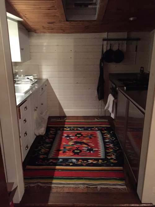 A cozy kitchen with wooden ceilings, white cabinets, and a colorful patterned rug on the floor.