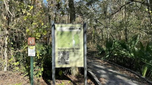 Sign for Bayou Coquille Trail in a wooded area, with a path leading into the greenery.