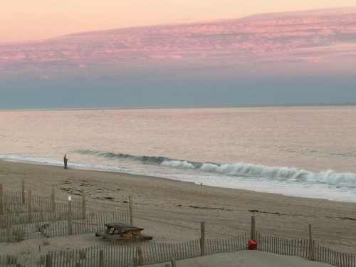 A serene beach at sunset, with gentle waves and a lone figure standing near the water's edge.