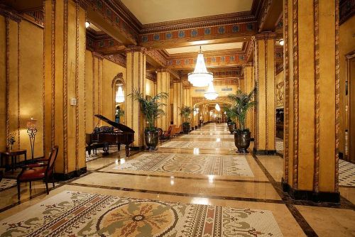 Elegant hotel lobby featuring ornate decor, tall columns, a grand piano, and lush plants under a chandelier.