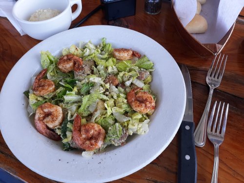 A plate of shrimp salad with lettuce, seasoning, and a side of rolls on a wooden table. Forks and a knife are nearby.