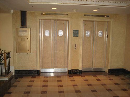 Two elevator doors with decorative designs in a hotel lobby, alongside a vintage mail slot on the wall.