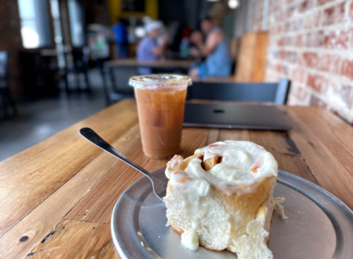 A cinnamon roll with cream cheese frosting on a plate, next to a cup of iced coffee in a cozy café setting.
