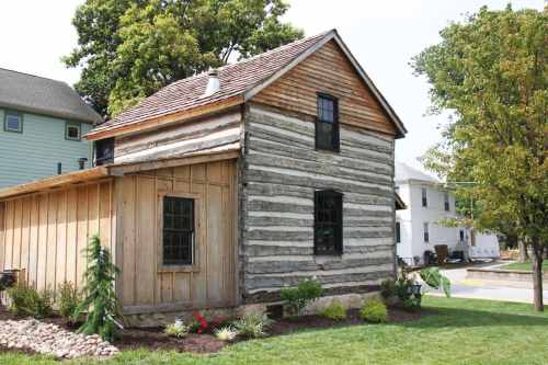 A rustic log cabin with a wooden addition, surrounded by greenery and flowers, set in a residential area.