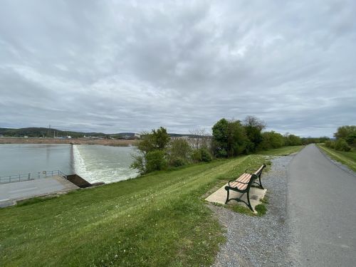 A serene riverside scene featuring a bench, grassy area, and a dam with water flowing, under a cloudy sky.