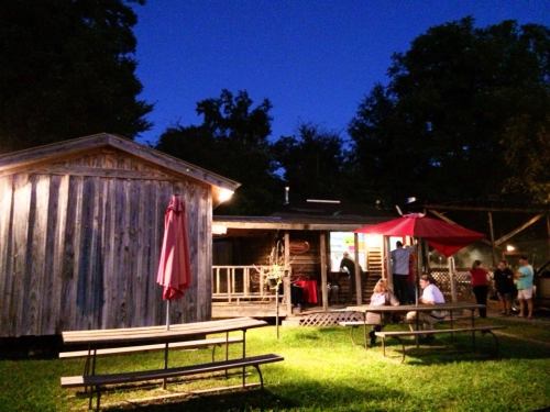 A rustic outdoor scene at dusk featuring a wooden building, picnic tables, and people gathered under red umbrellas.
