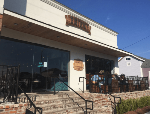 A modern restaurant exterior with a wooden sign, large windows, and outdoor seating on a sunny day.