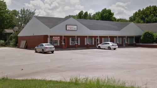 A single-story brick building with a sign, surrounded by a parking lot and greenery under a cloudy sky.