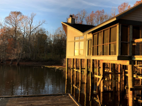 A lakeside house on stilts, reflecting sunlight, surrounded by trees with autumn foliage.
