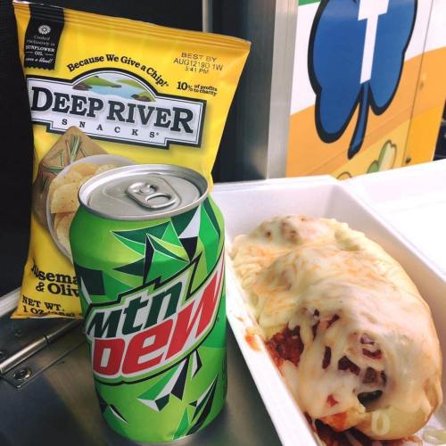 A can of Mountain Dew next to a plate of cheesy food and a bag of Deep River Snacks on a table.