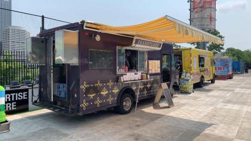 A food truck with a yellow awning parked beside another truck, set in an outdoor area with buildings in the background.