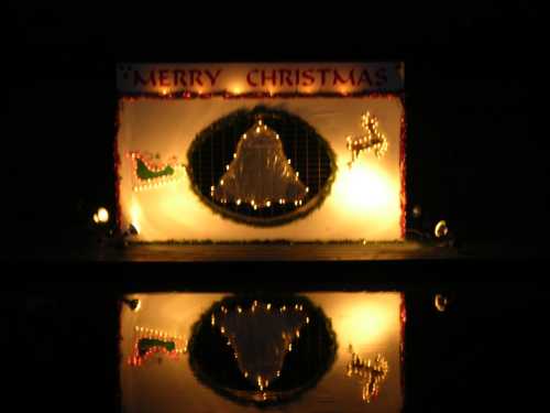 A festive display with "Merry Christmas," a bell, and lights, reflecting on water at night.