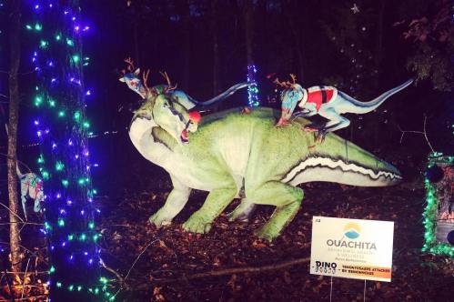 A festive dinosaur display with colorful lights, featuring dinosaurs in holiday attire and reindeer antlers.