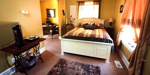 Cozy bedroom with a large bed, warm yellow walls, and a small sitting area in the background. Soft lighting and decor.