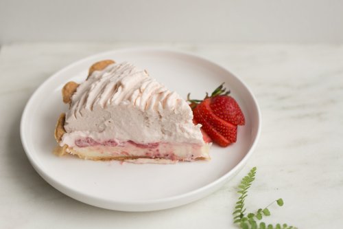A slice of strawberry cream pie on a white plate, garnished with fresh strawberries and a sprig of fern.