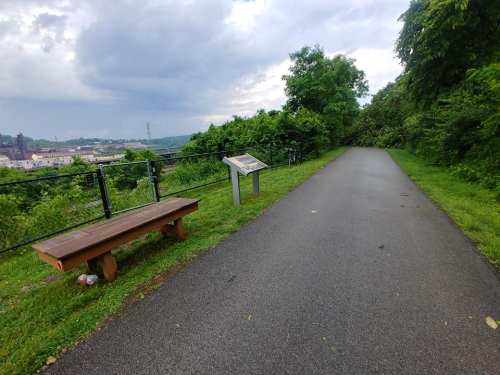 A scenic pathway lined with greenery, featuring a wooden bench and an informational sign.