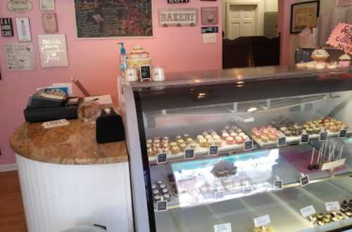 A cozy bakery interior with a pink wall, a display case filled with colorful cupcakes, and a counter with decor.