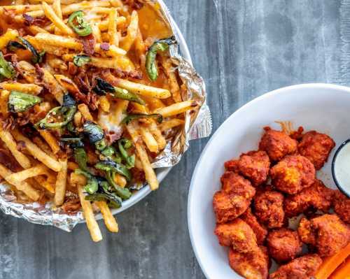 A bowl of spicy buffalo chicken bites with a side of ranch dressing, next to a tray of loaded fries topped with cheese and peppers.