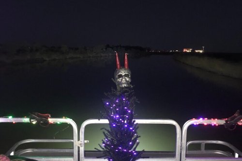 A decorated boat at night features a skull with horns and a lit Christmas tree, reflecting on the water.