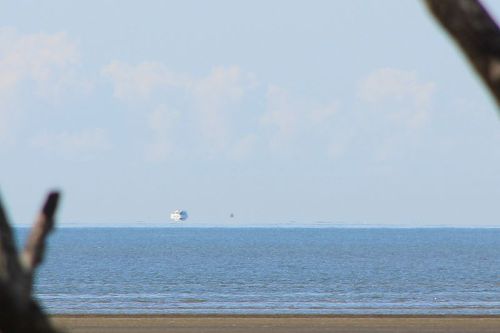 A distant ship and a small sailboat on a calm blue sea, with a clear sky and soft clouds in the background.