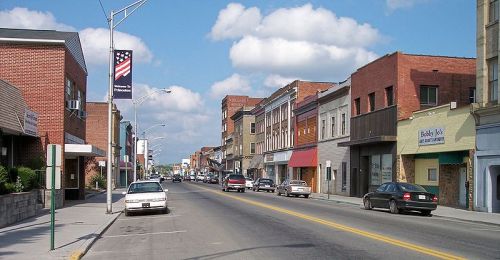 A street view of a small town with brick buildings, shops, and parked cars under a blue sky with fluffy clouds.