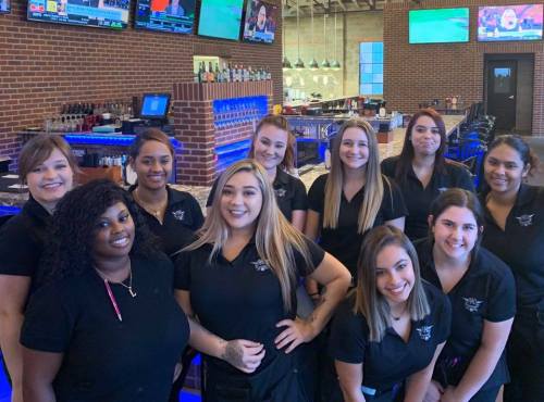 A group of ten women in black shirts smiling together in a bar setting with a blue-lit counter and TVs in the background.