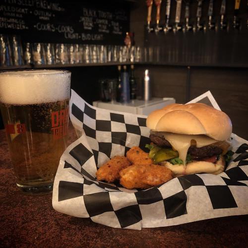 A glass of beer next to a plate of a burger with cheese, pickles, and a side of crispy chicken bites on a checkered paper.