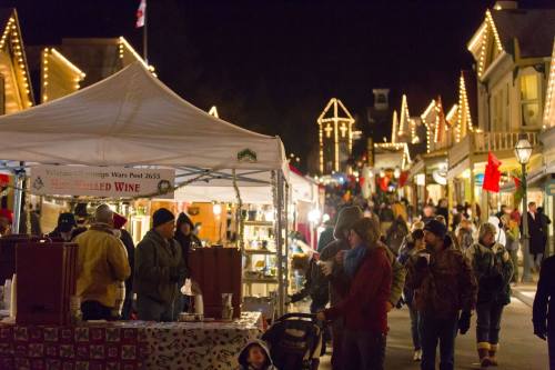 A festive street market at night, with people browsing stalls and holiday lights illuminating the scene.