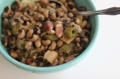 A turquoise bowl filled with black-eyed peas, green peppers, and diced bacon, with a spoon resting on the side.