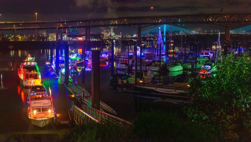 Colorful boats illuminated at night in a marina, with reflections on the water and a bridge in the background.