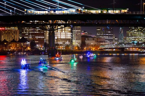 Colorful boats with lights navigate a river at night, under a bridge with city buildings illuminated in the background.