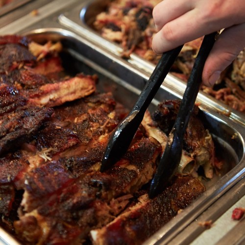 A hand using tongs to serve tender, smoky barbecue ribs from a metal tray.