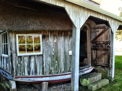 A rustic wooden building with a thatched roof, featuring a small window and an open door, set in a grassy area.